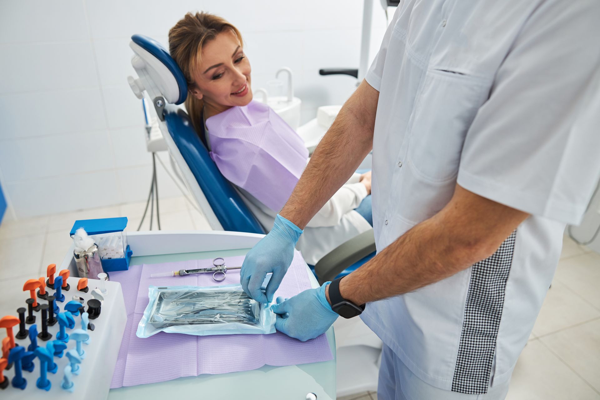 Dentist preparing dental instruments beside a reclining patient in a bright clinic
