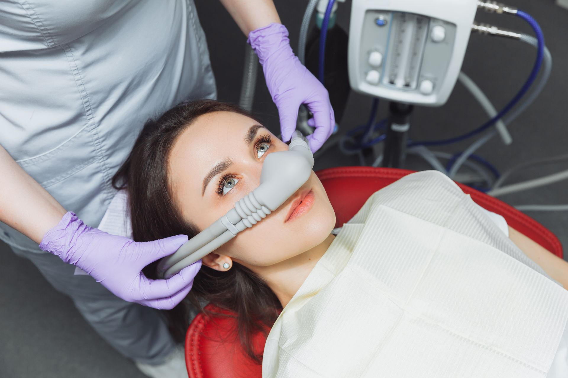A dental professional in gloves places a nitrous oxide sedation mask over a patient's nose in a clinical setting.
