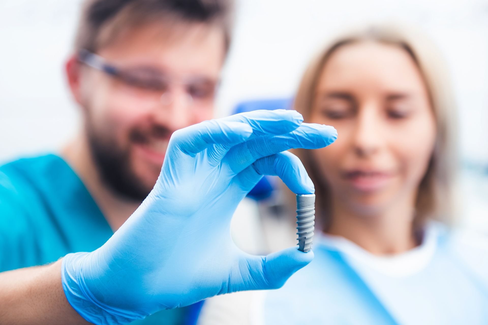 A person in a medical uniform and blue gloves holds a small dental implant, with another person visible in the background.