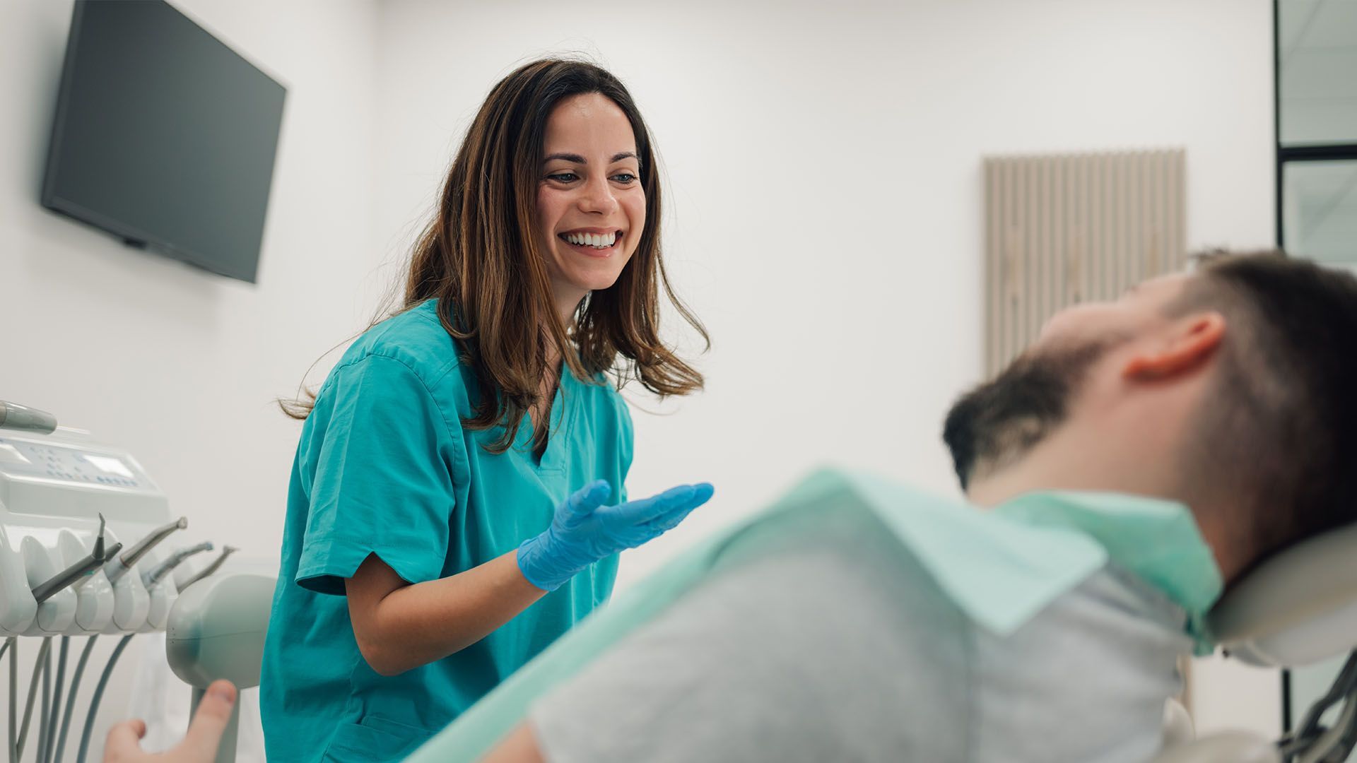 Dentist in teal scrubs smiles at patient in dental chair, dental office setting.