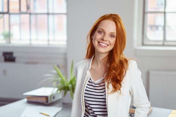 A woman with red hair is smiling while sitting at a desk in an office.