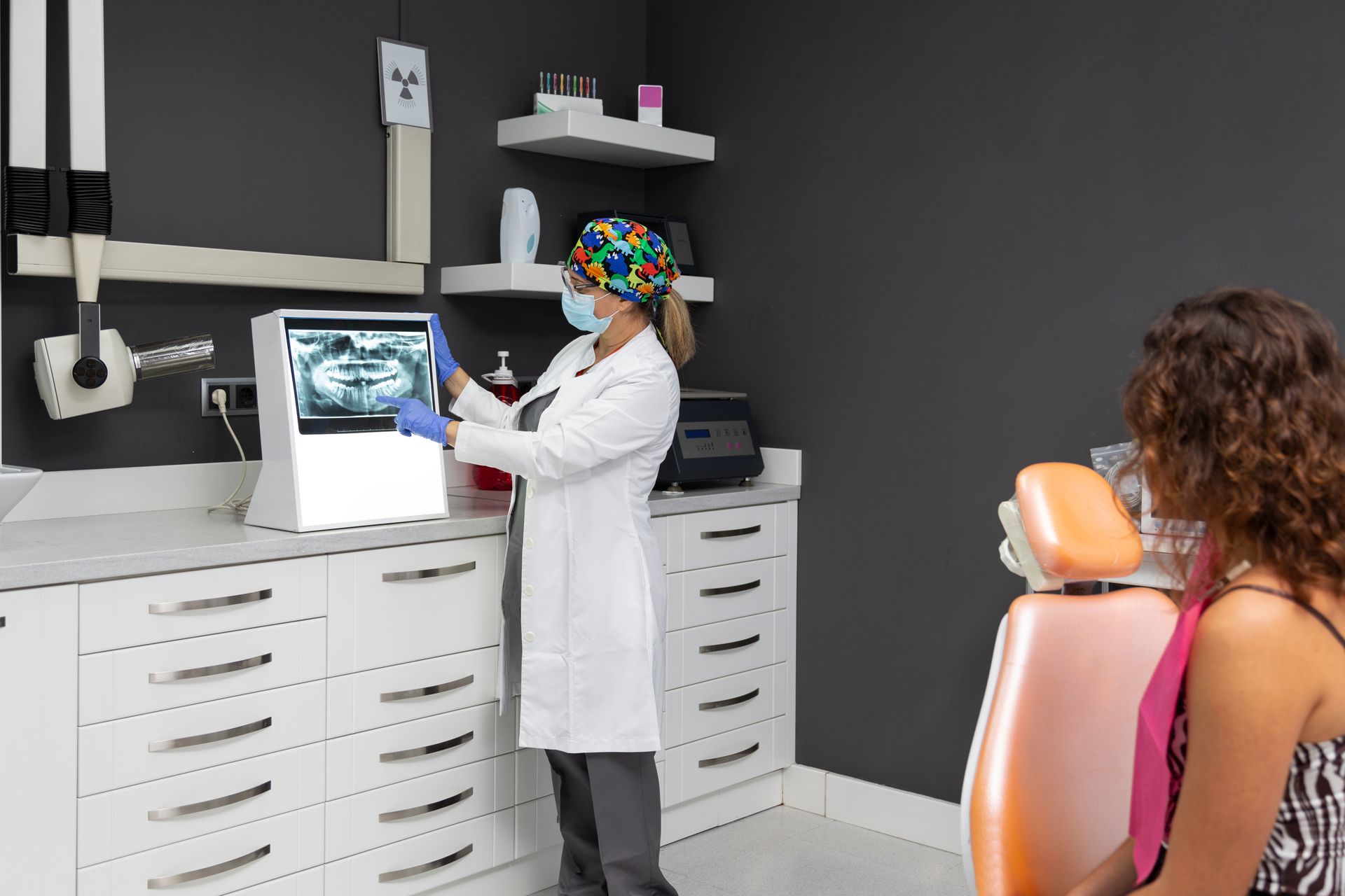 A dentist in a white coat and mask points to a panoramic dental X-ray on a computer screen for a patient in a dental chair.