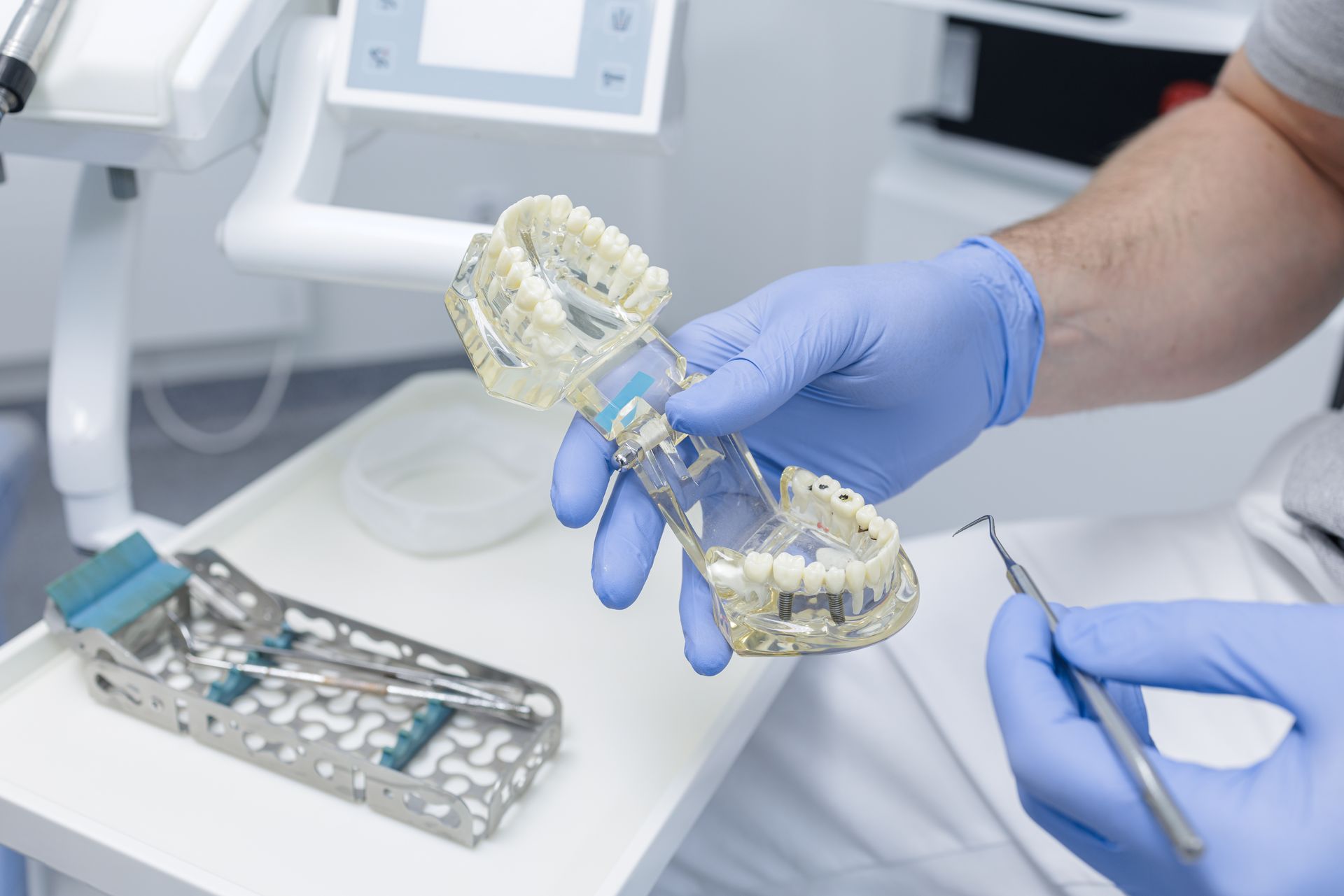 Dentist holding a jaw model in blue gloves, with dental tools on a tray in a clinic.