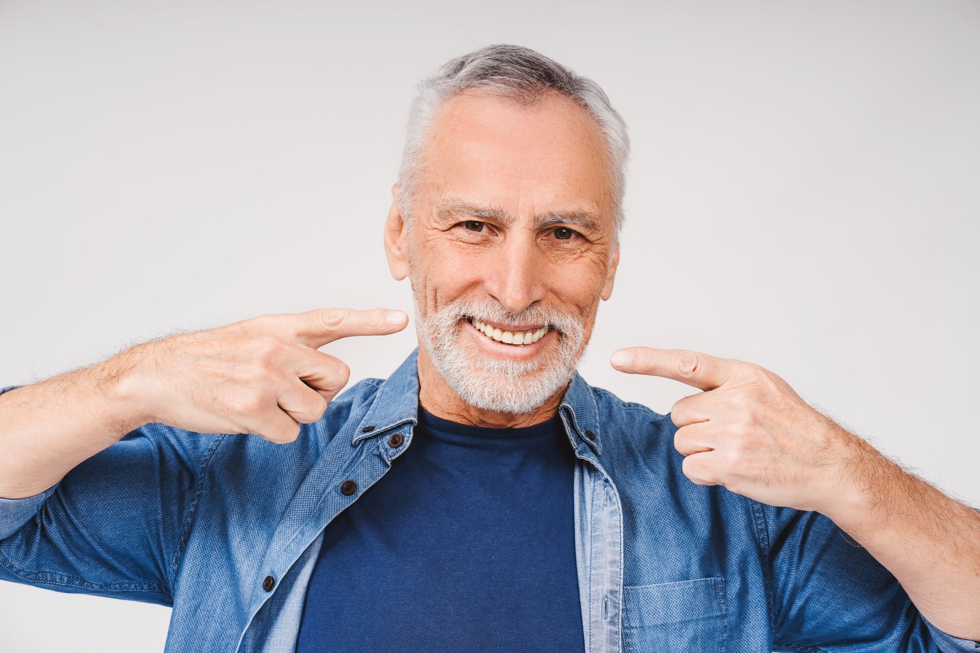 Man in blue shirt smiles, pointing at his teeth. Gray hair, light background.
