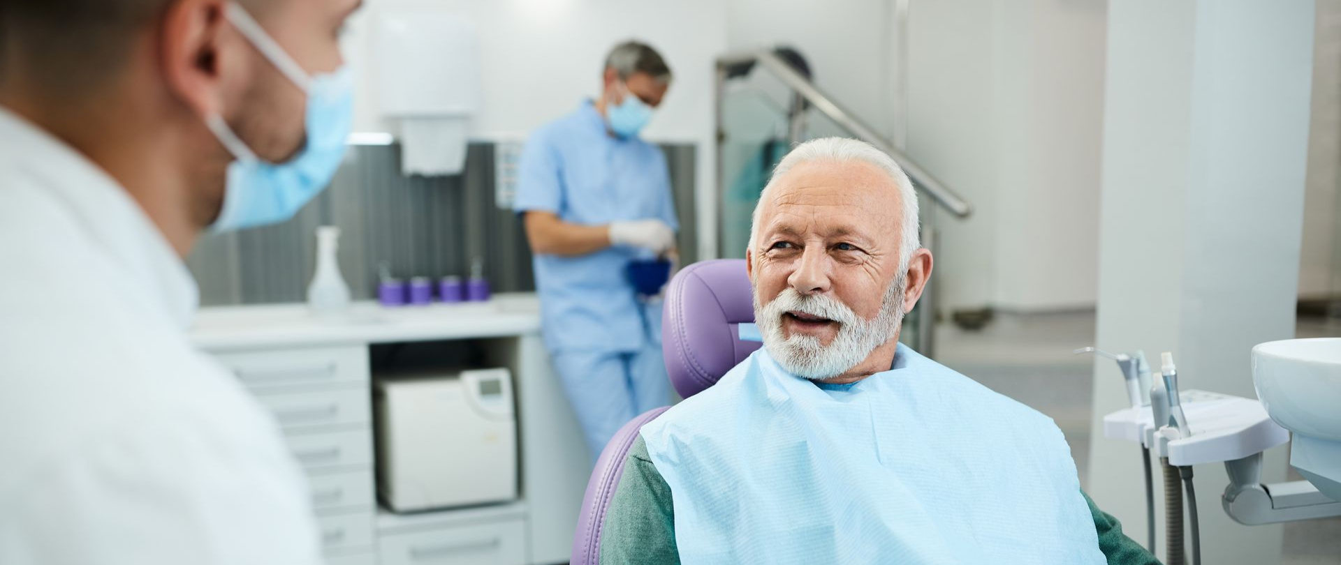 Elderly man at dentist, facing dentist. Dental assistant in background.