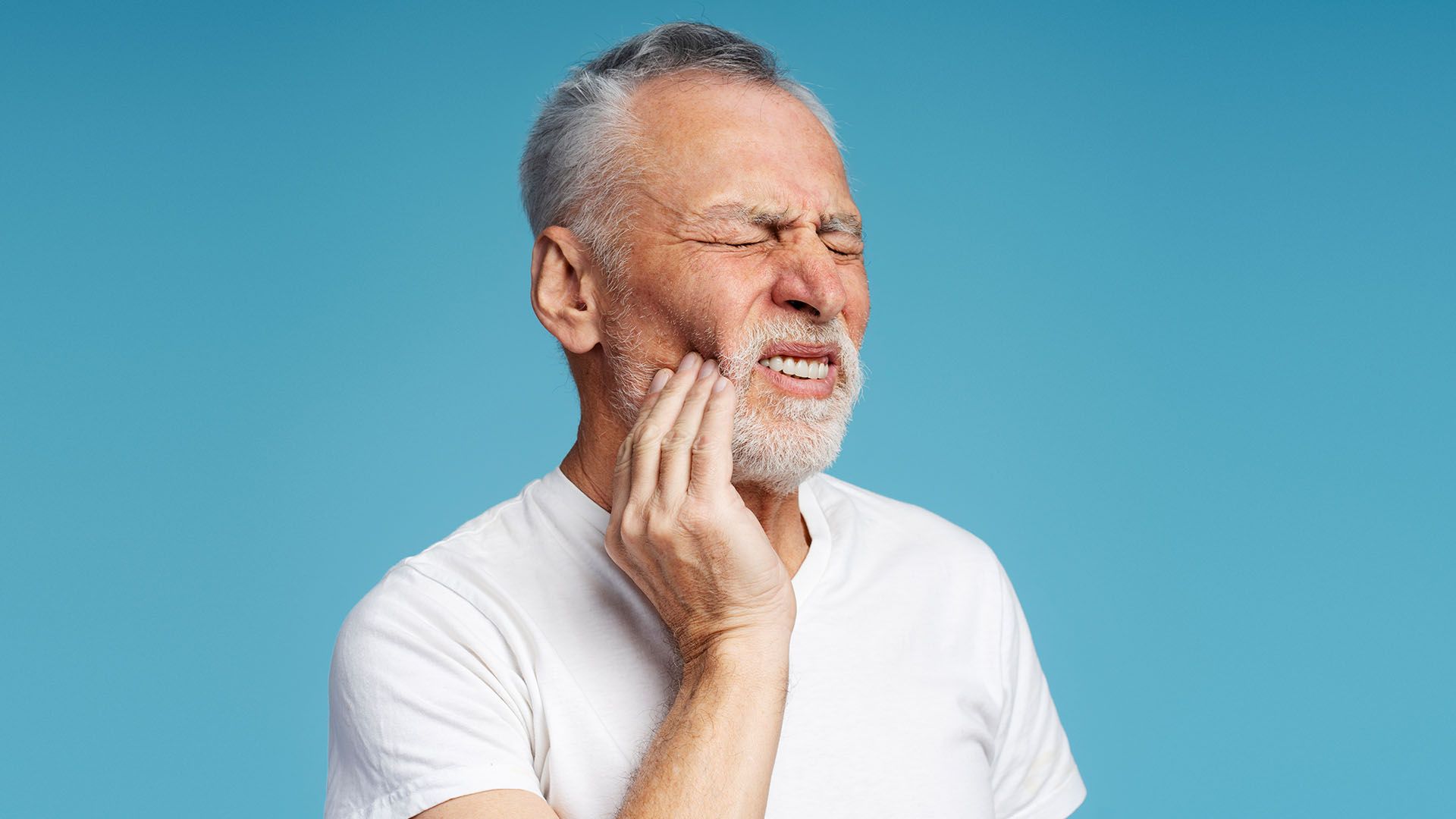 An older man, earing a white shirt, clutches his jaw with a pained expression
