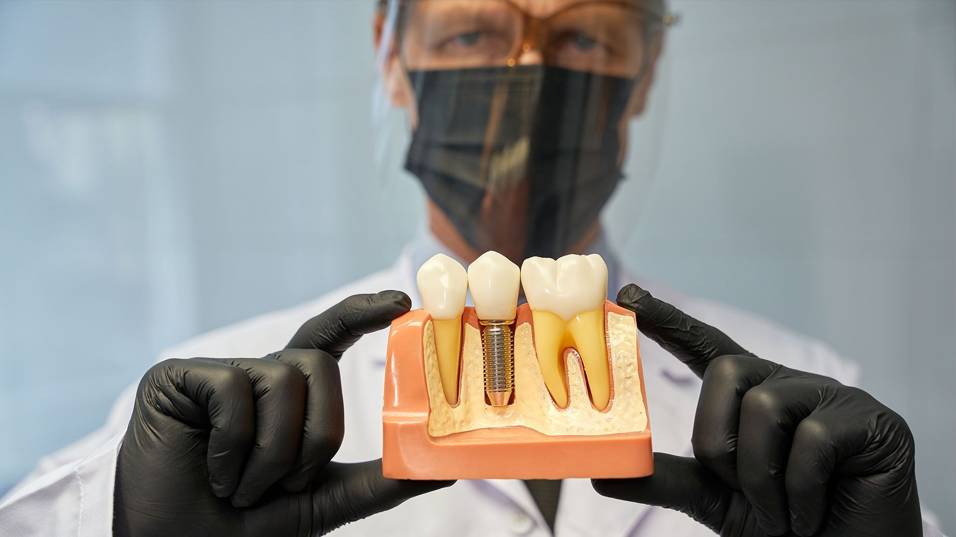 A dentist wearing a mask and gloves holds a model of teeth, including a dental implant.