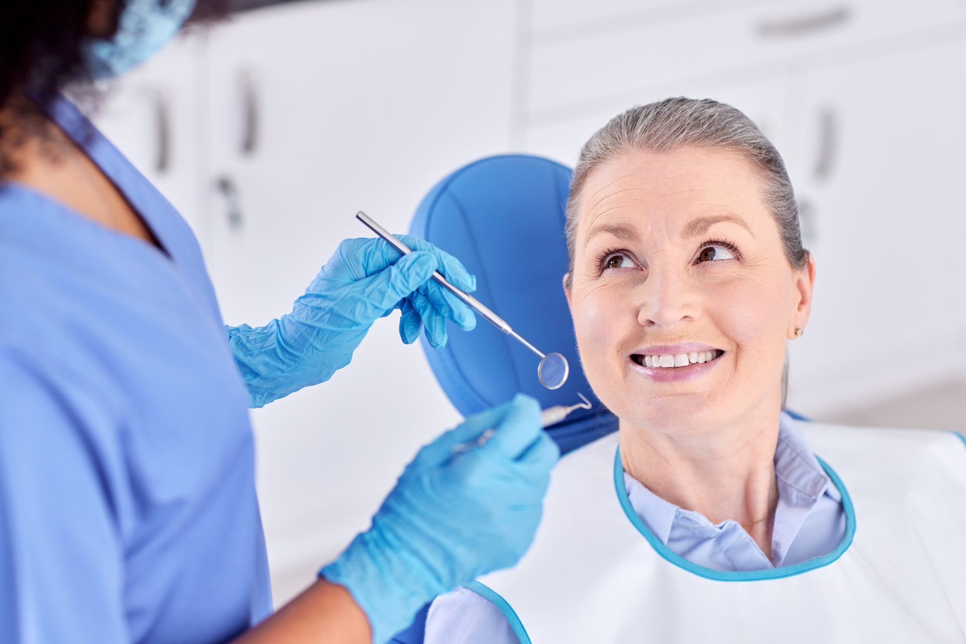 Dentist examining a patient's teeth. The patient smiles, wearing a bib, seated in a dental chair.