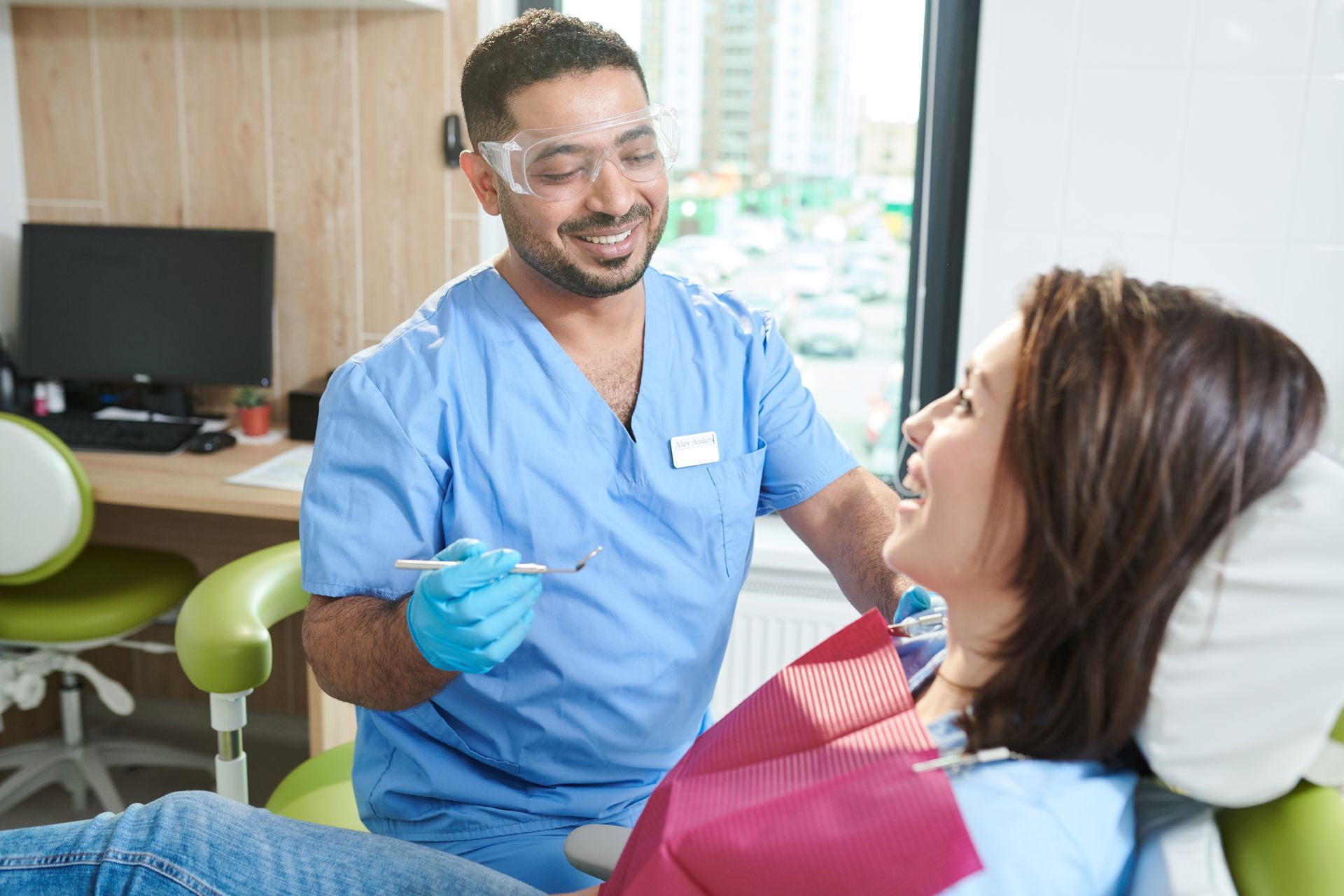 Dentist in blue scrubs examining a woman's teeth in a dental office. Both are smiling.