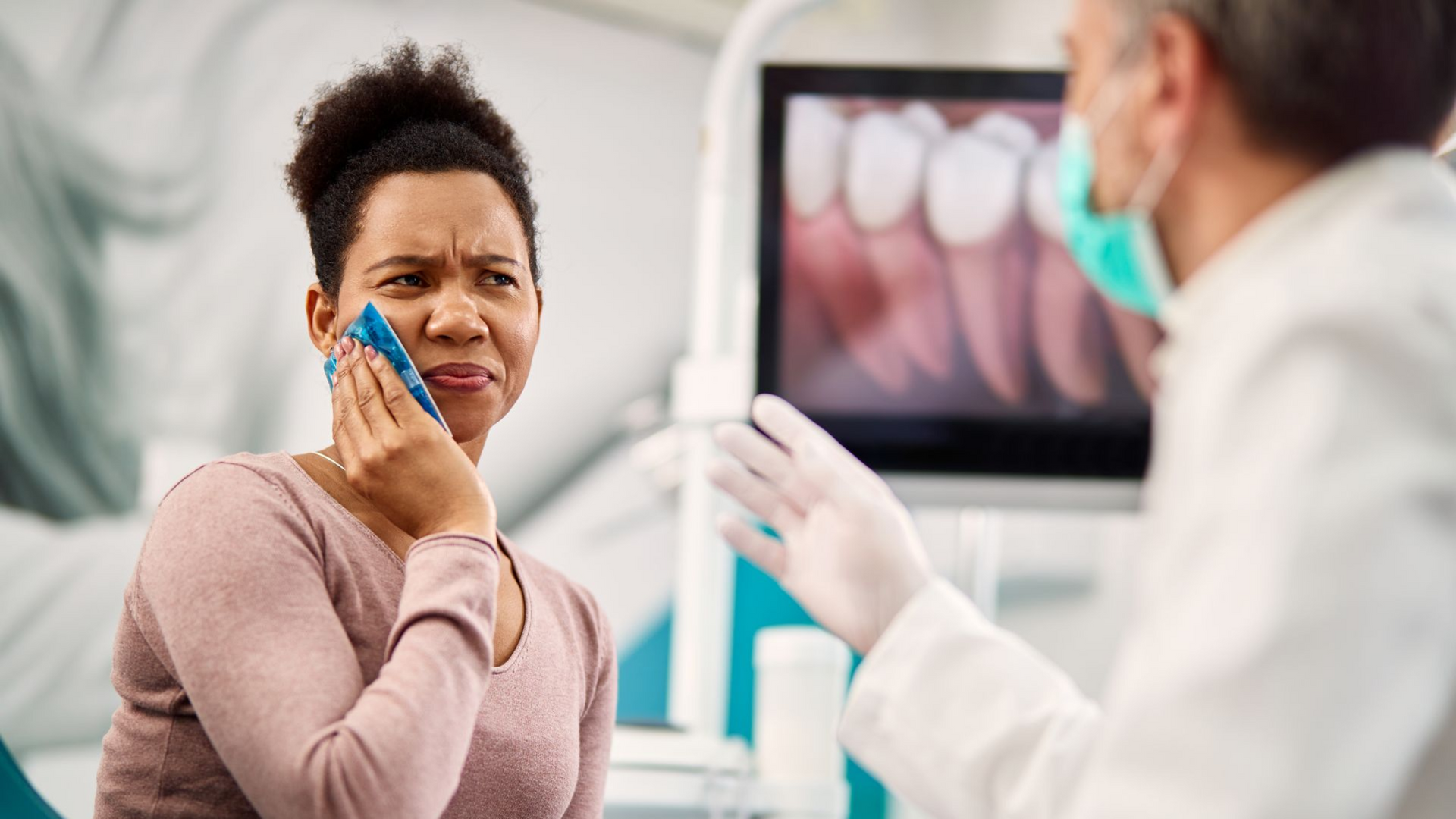Woman at dentist, holding jaw, in pain. Dentist pointing at screen with teeth.