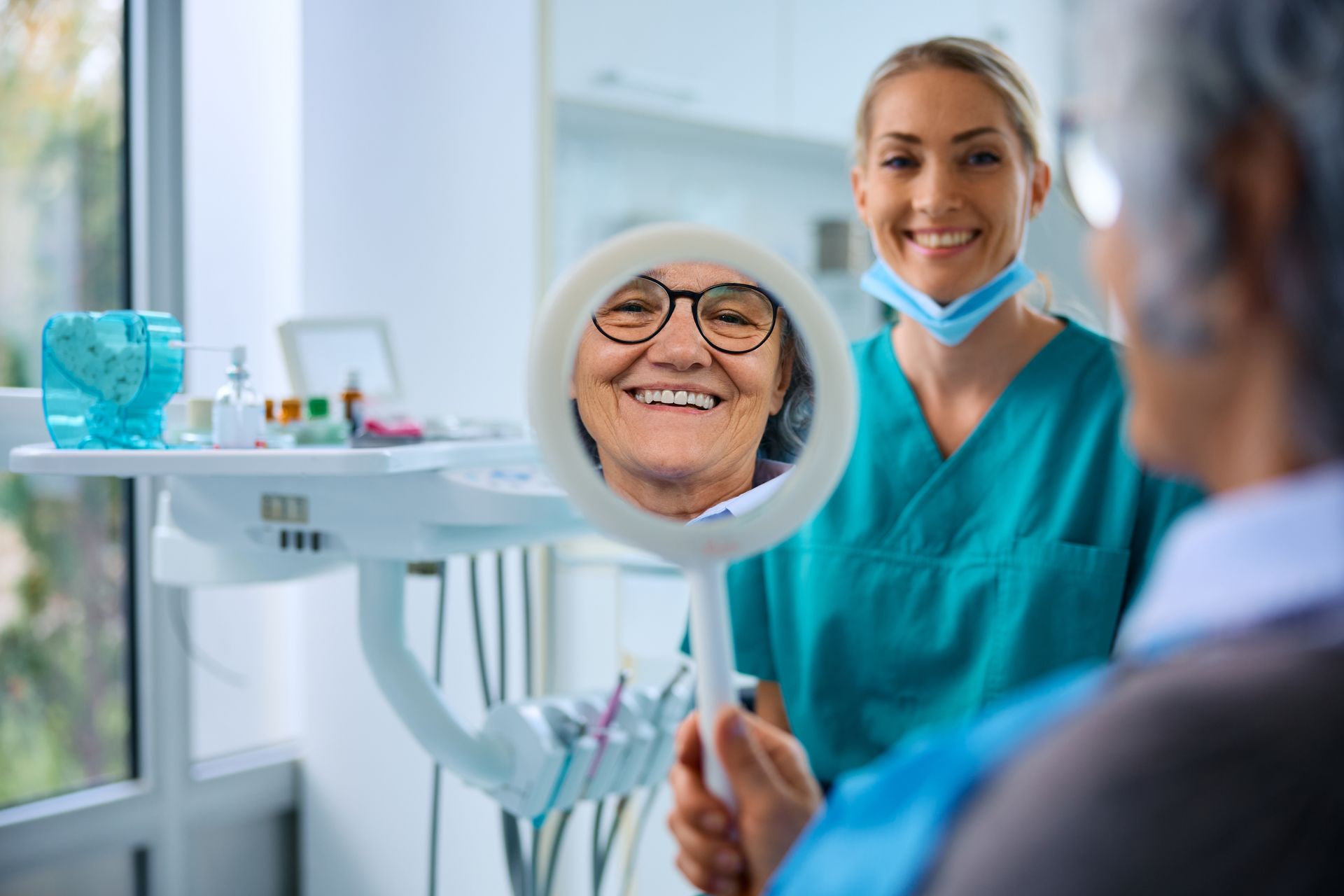 Dentist holds mirror for smiling patient, showing teeth. Dentist in green scrubs, office setting.