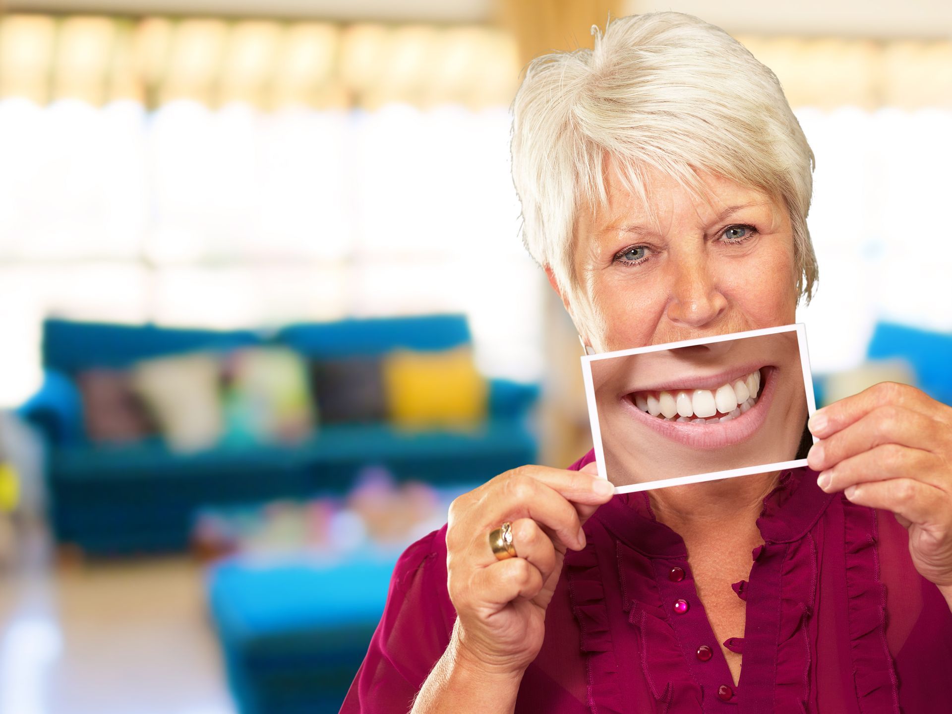Woman holding a photo of teeth over her mouth, smiling. Living room in background.