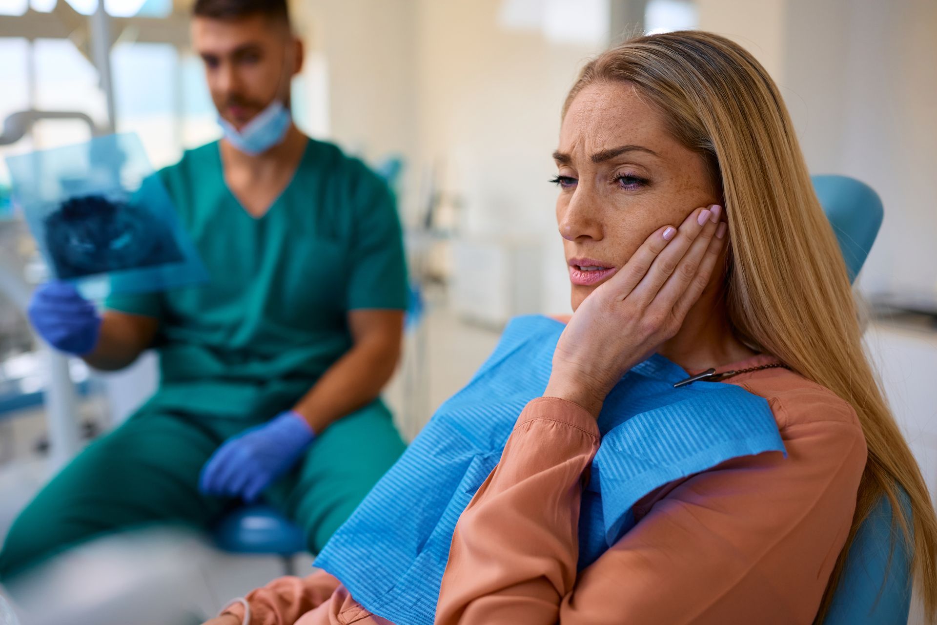 Concerned patient in blue dental chair, hand on cheek, with dentist blurred in background