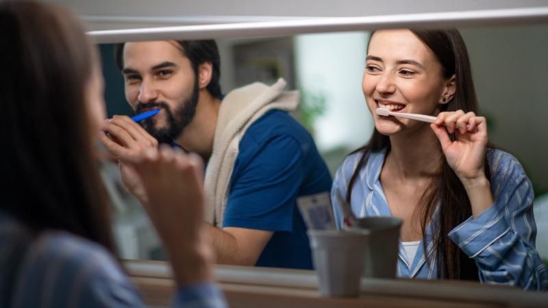 A man and a woman are brushing their teeth in front of a mirror.