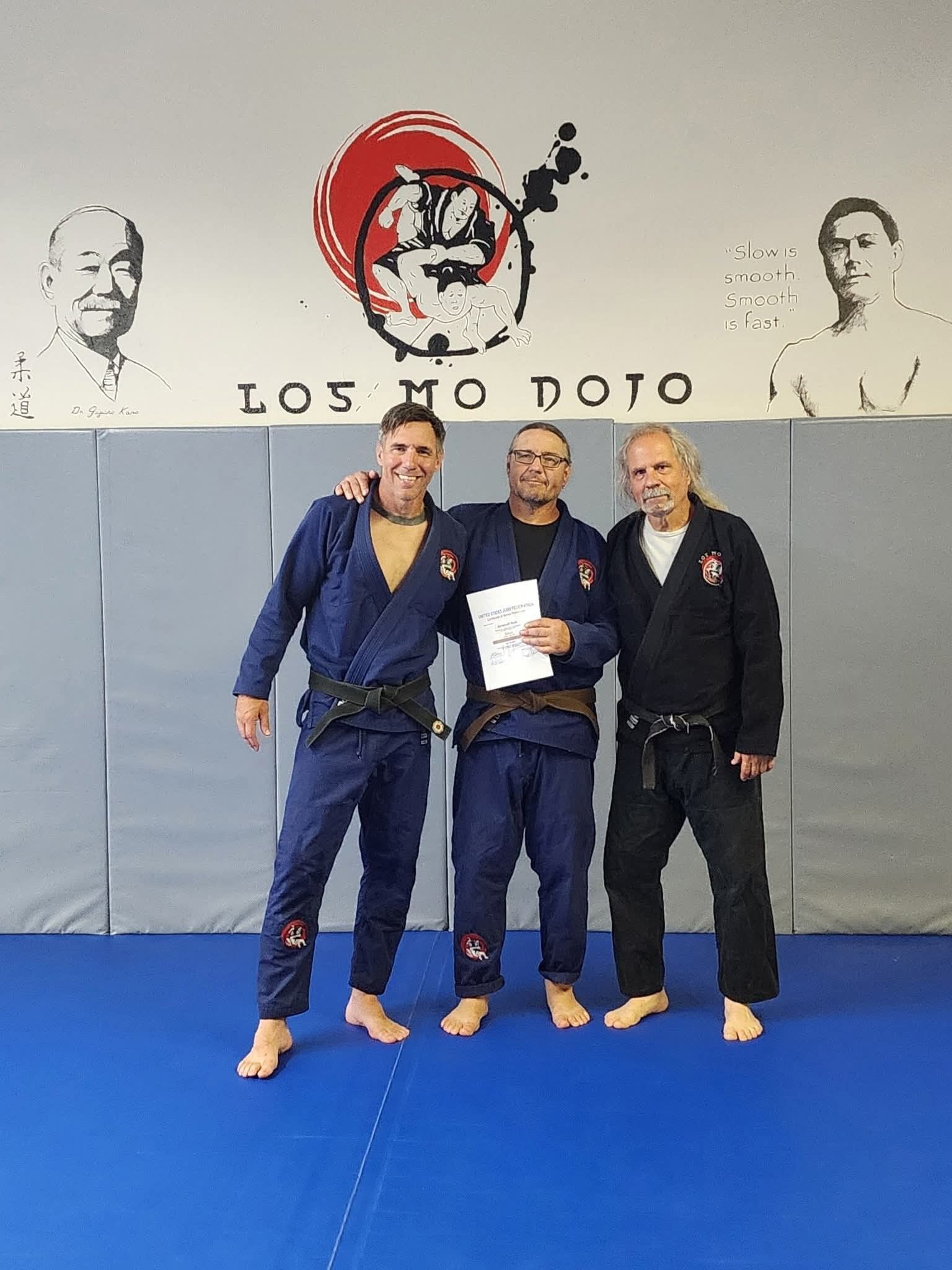 Three men in martial arts uniforms stand on a blue mat in front of a wall at Los Mo Dojo. Two are in blue gis, one holds a certificate.
