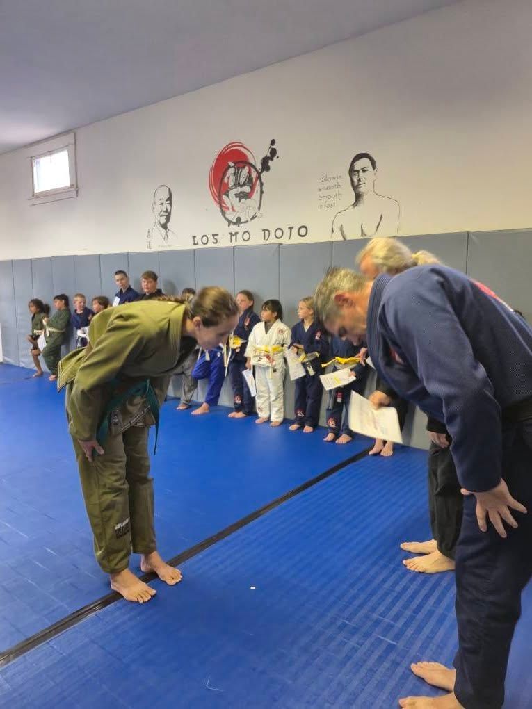 People bowing at a Jiu-Jitsu promotion ceremony on a blue mat zt Los Mo Dojo. Others in gis and belts stand behind them.
