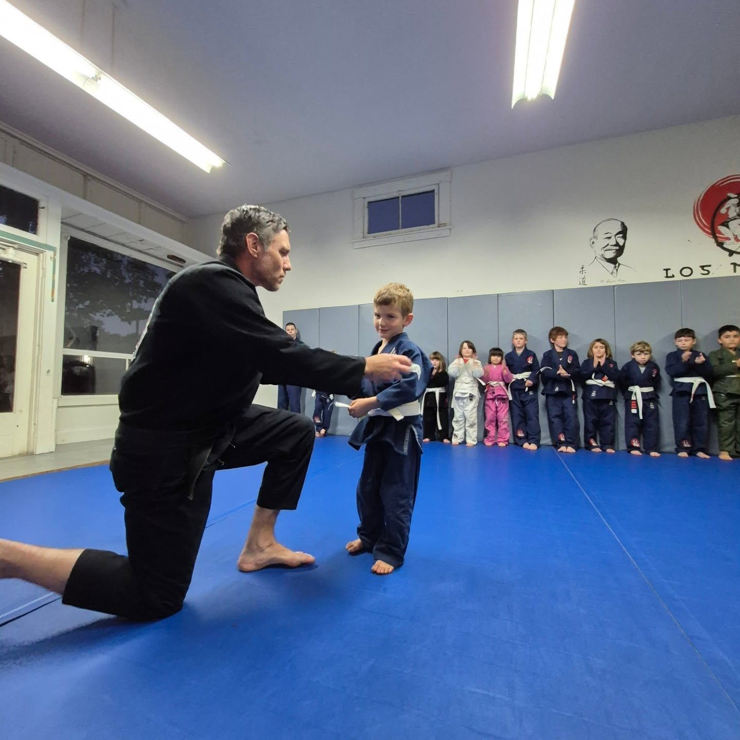A man kneeling, adjusting the belt of a child in a martial arts class at Los Mo Dojo. Other children watch.