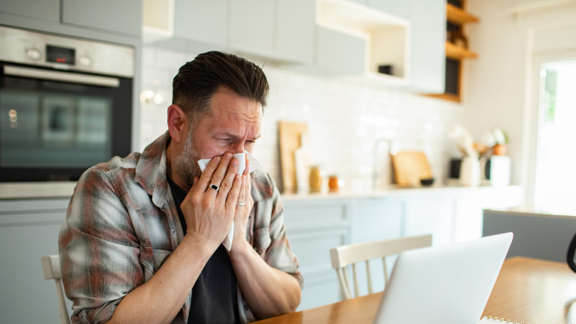 Man blowing his nose with a tissue while sitting at a table with a laptop in a kitchen setting.