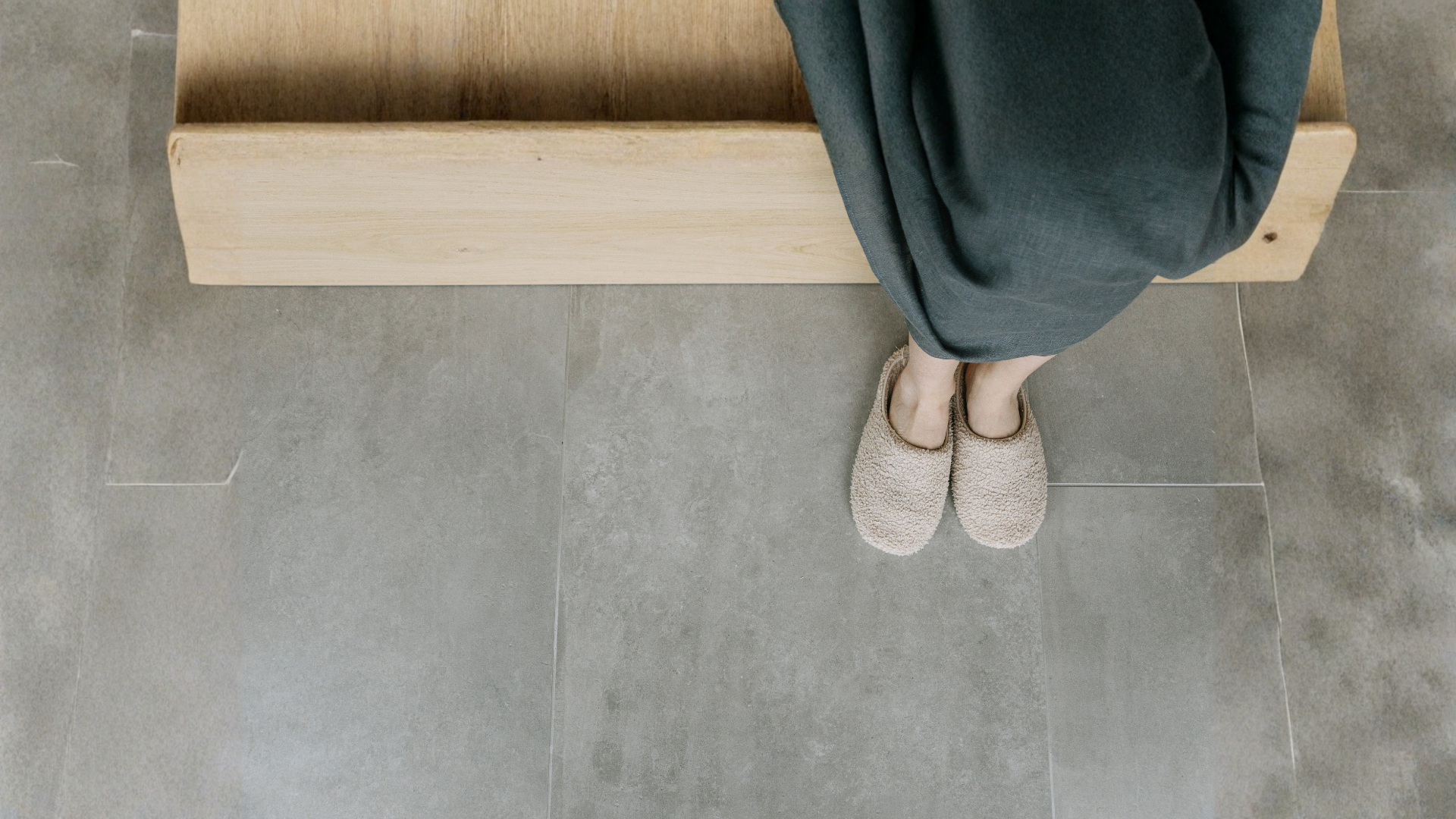 Person seated on wooden bench, feet in fuzzy slippers, gray floor.