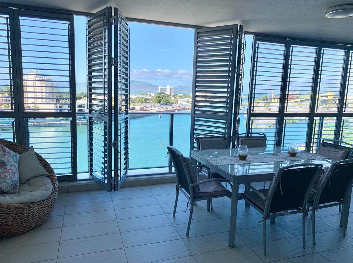 Balcony with ocean view, folding shutter doors, outdoor dining table, wicker chair, blue water, sunny day.