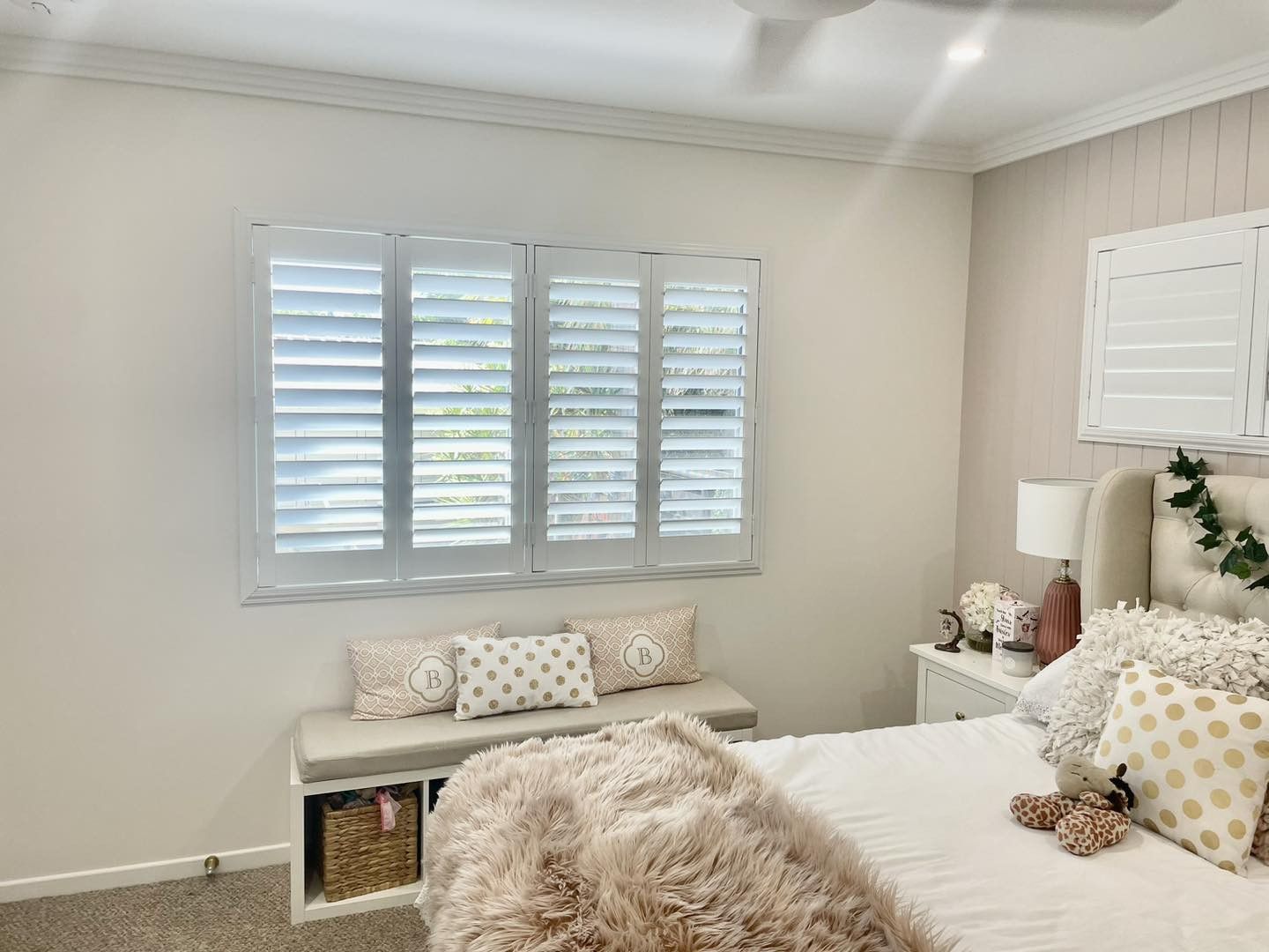 Bedroom with closed white shutters, bench with pillows, bed with decorative pillows and a nightstand.
