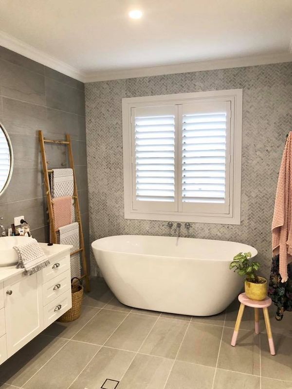 Modern bathroom with a white freestanding tub, shuttered window, gray tile, and wooden ladder with towels.