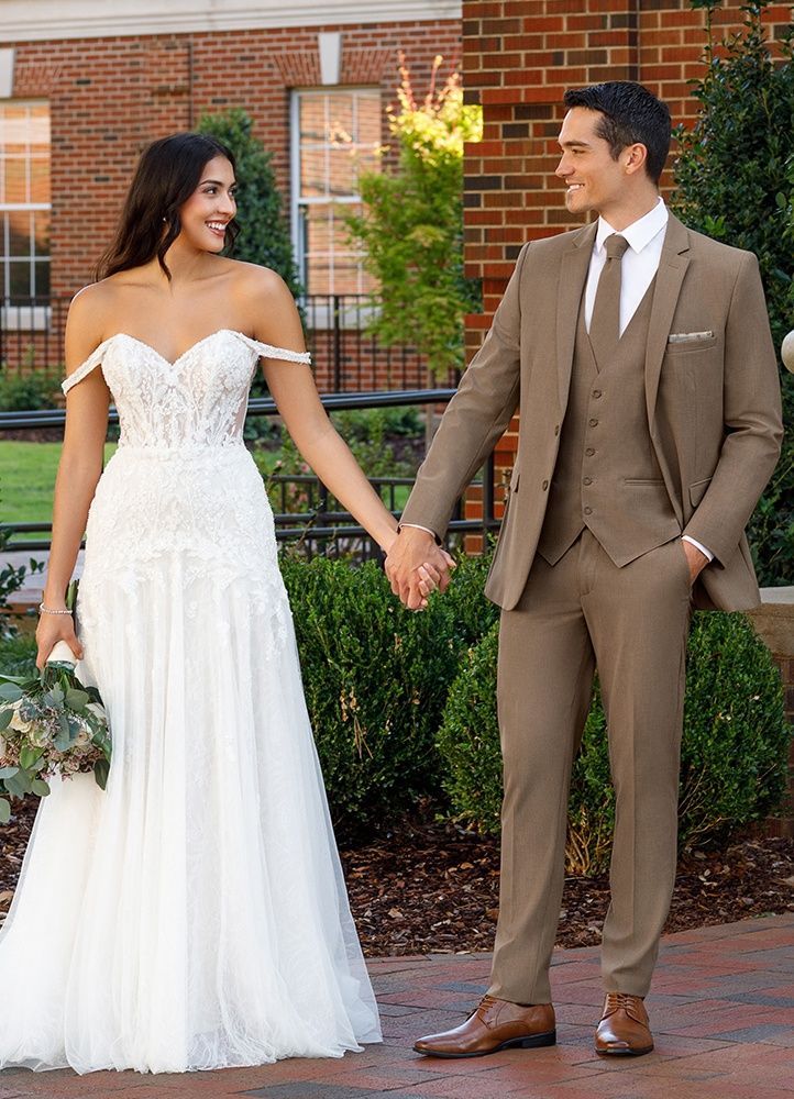 Bride and groom holding hands, smiling. The bride wears a white gown, and the groom a brown suit. Outdoors.