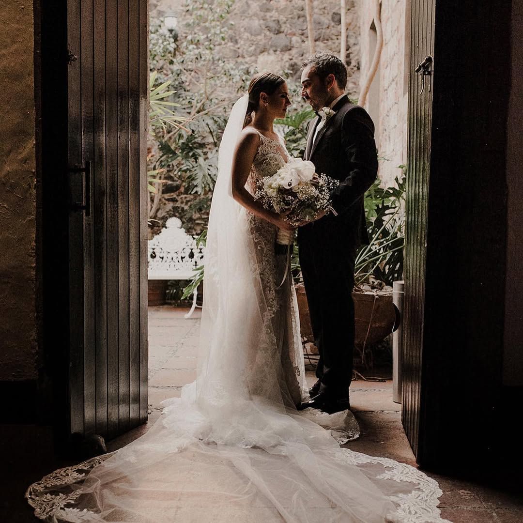 Couple in wedding attire facing each other, standing in a doorway with floral arrangements and greenery in background.