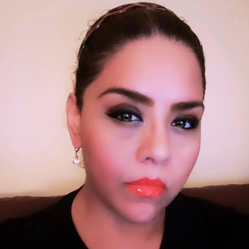 A woman with dark hair and dramatic eye makeup wears a black top, hoop earrings, and orange lipstick.