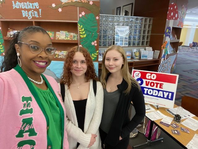 Three females are posing for a picture in front of a sign that says go vote today