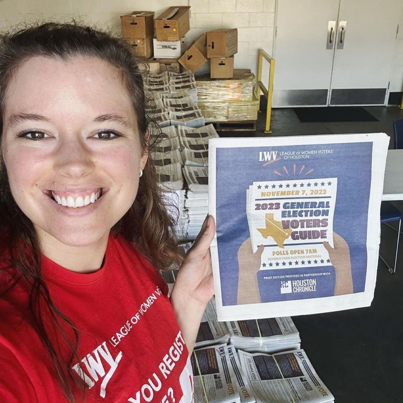 A woman wearing a red shirt that says you register holds up a newspaper