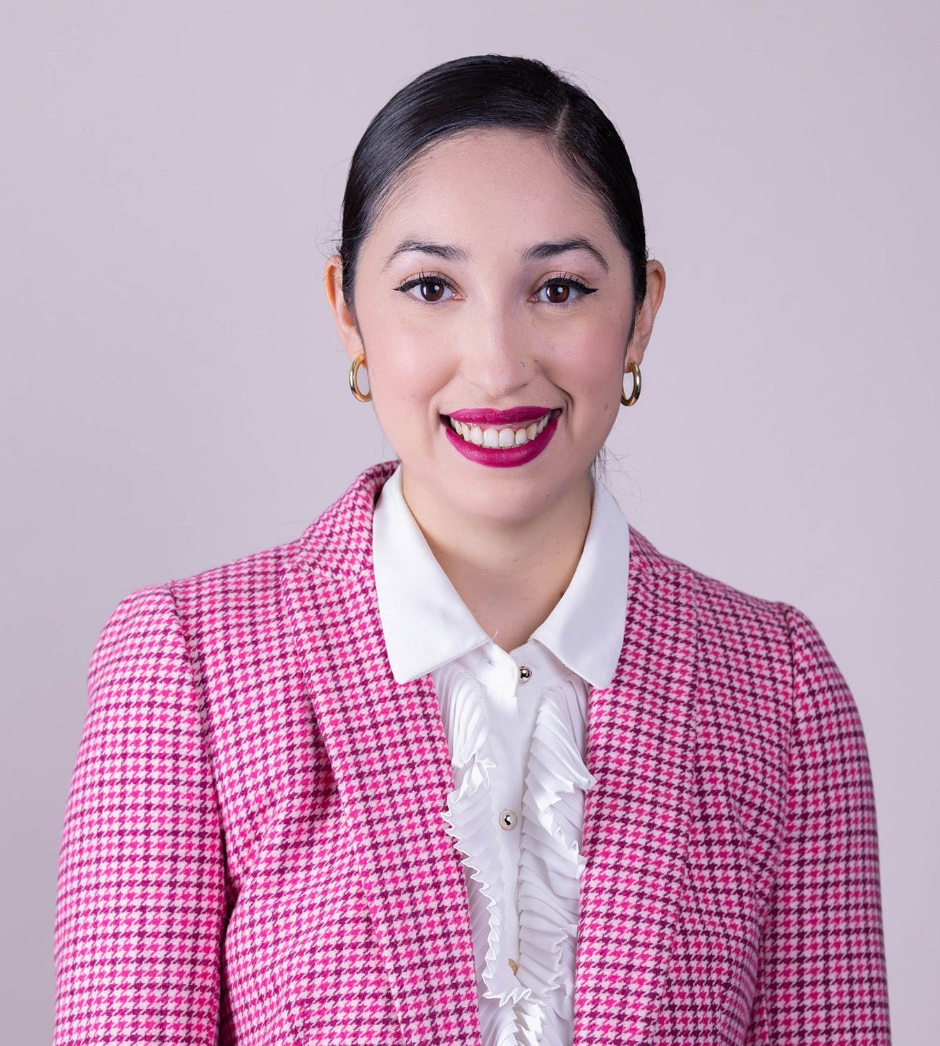 Woman smiling, wearing a pink blazer, white ruffled shirt, and gold earrings. Light pink background.