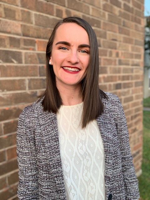 Woman with shoulder-length brown hair smiles in front of a brick wall, wearing a gray and white tweed blazer and a white sweater.