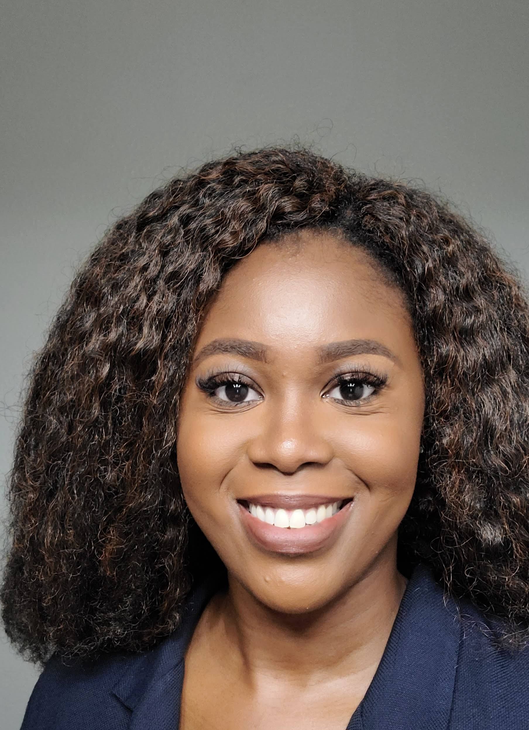 A smiling Black woman with dark, curly hair wearing a navy blazer. She has a neutral background.
