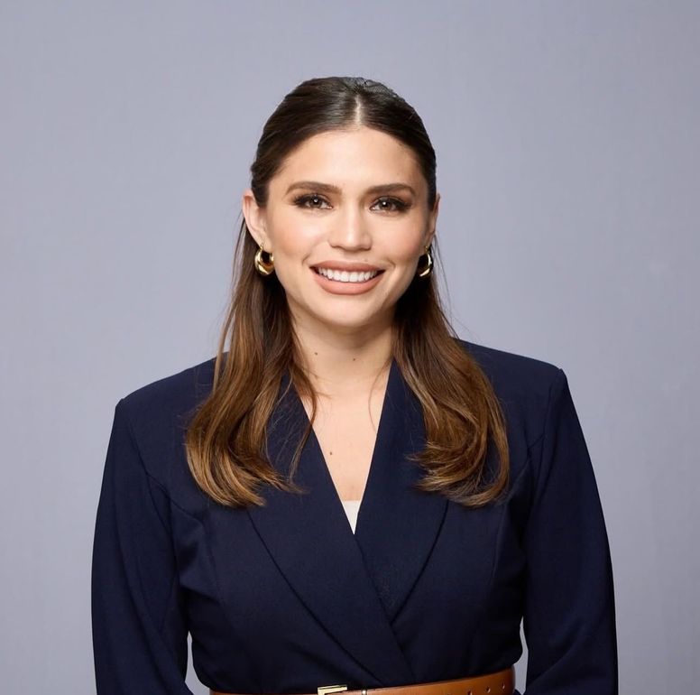 A woman wearing a green jacket and a pearl necklace is smiling for the camera.
