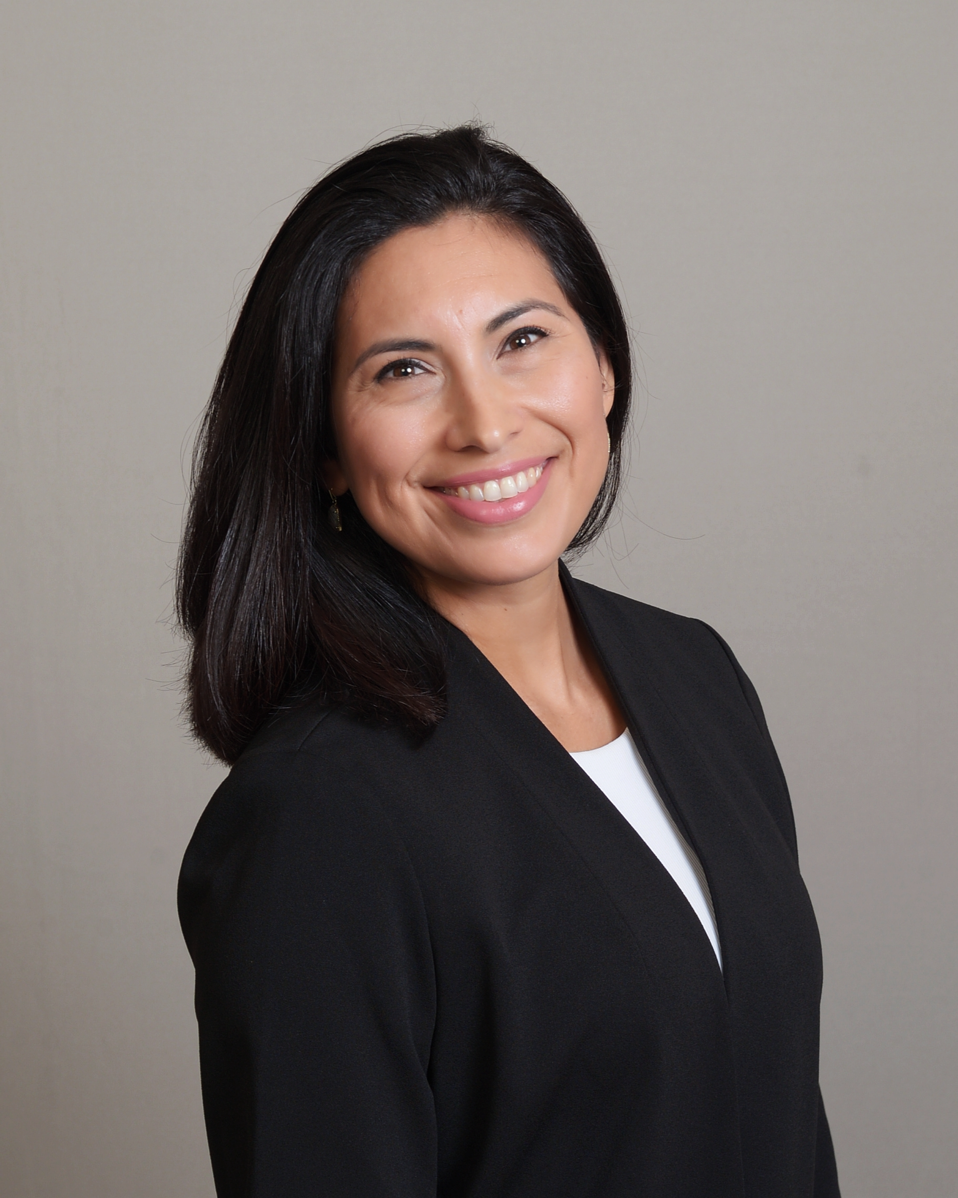 Woman with dark hair smiles, wearing a black blazer over a white shirt, set against a gray background.