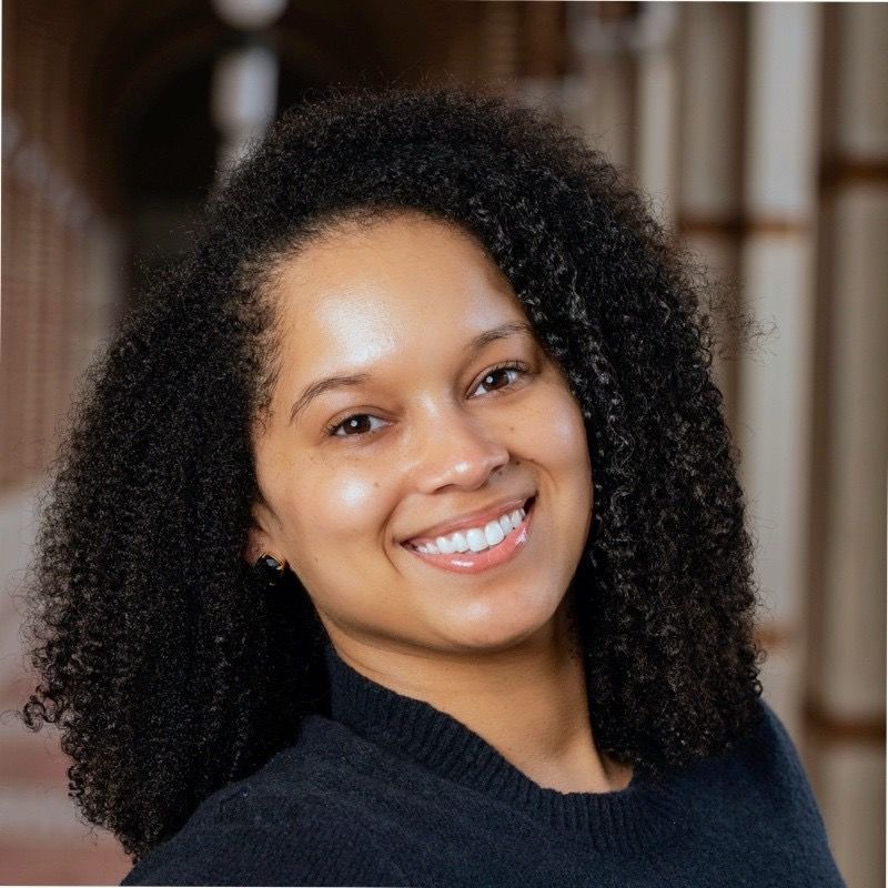 Smiling woman with dark curly hair, wearing a black sweater. She is indoors, likely a building hallway.