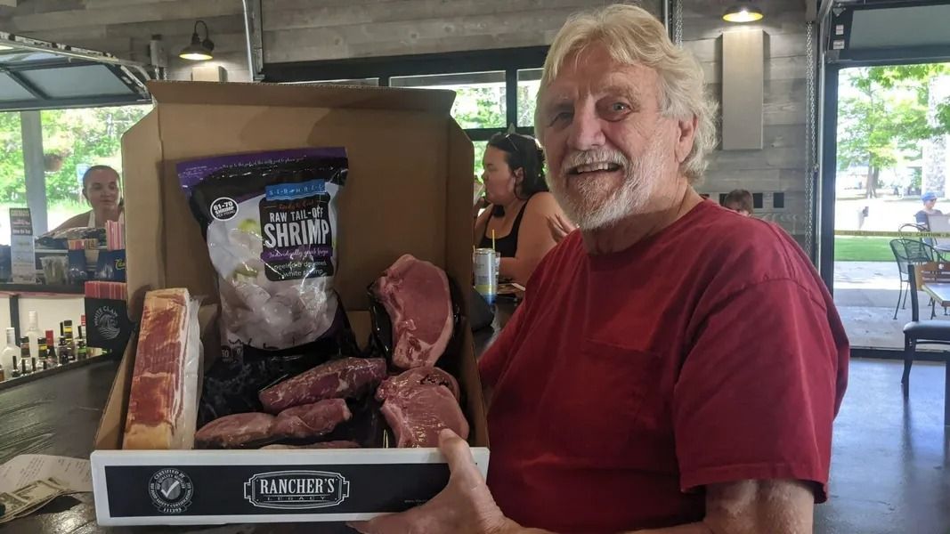 Smiling man in a red shirt holding a display of smoked meats at an indoor market booth