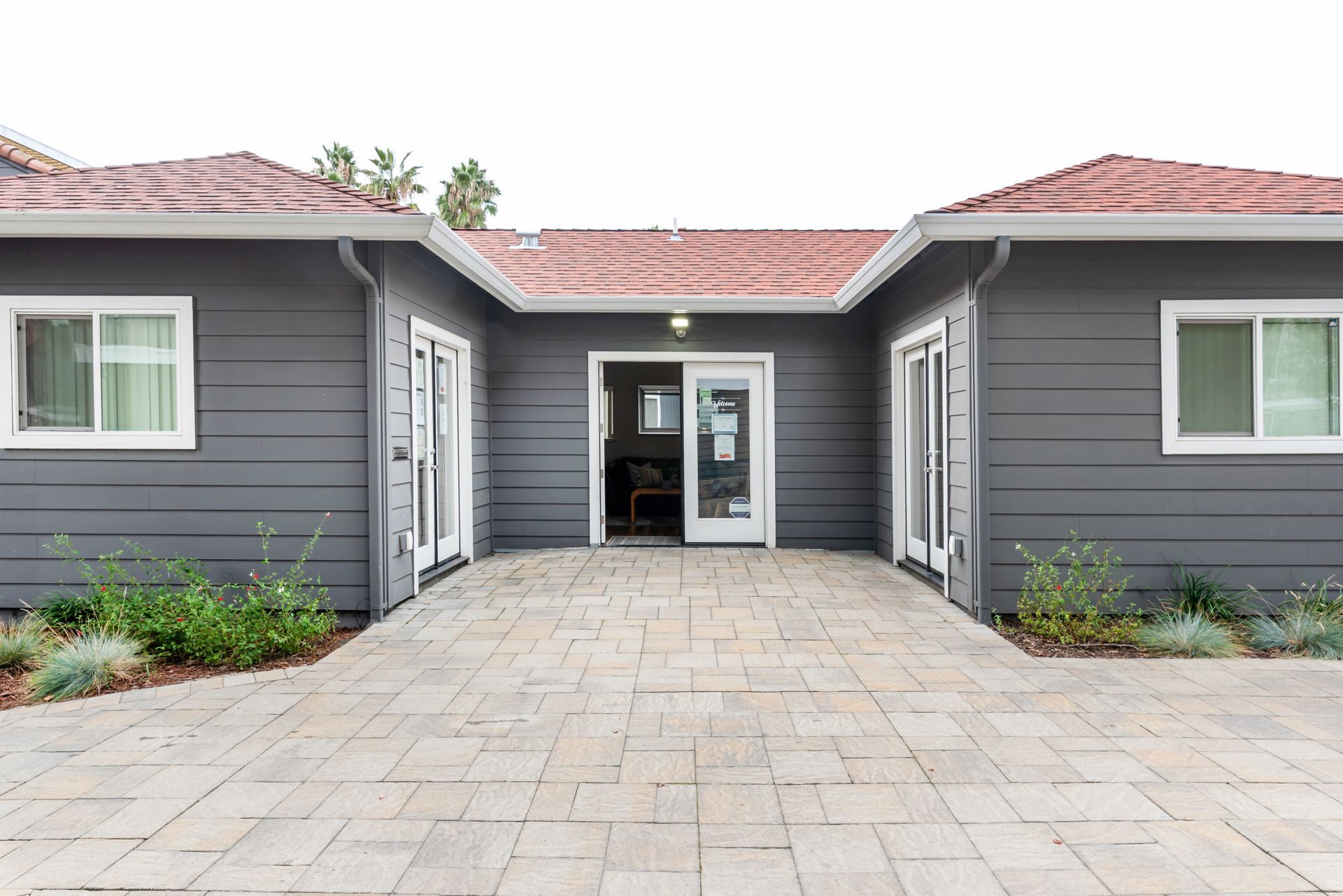 Exterior of a gray building with red tile roof, glass doors and windows, and a brick pathway.