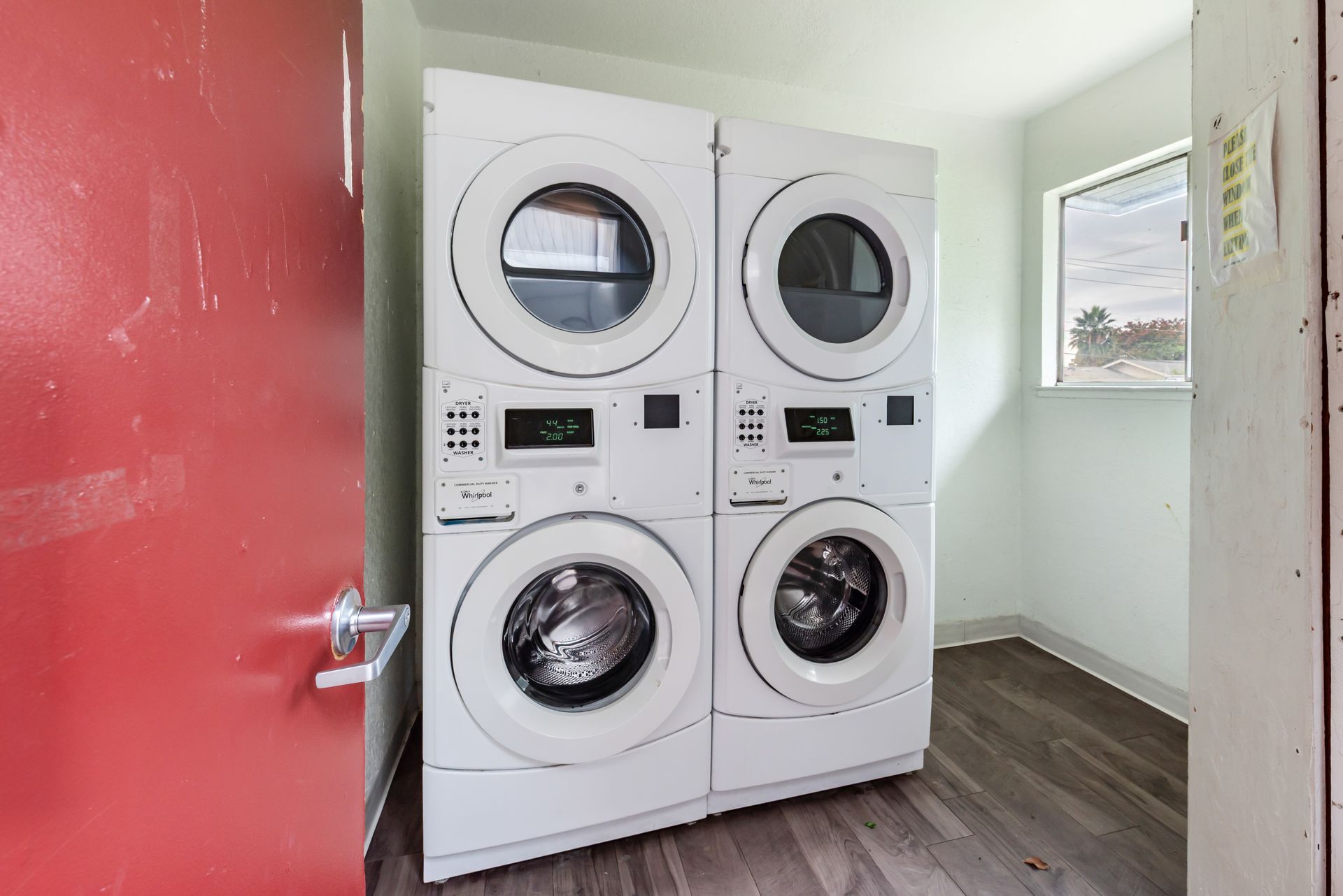 Stacked white washer and dryer units in a laundry room, next to a red door and a small window.