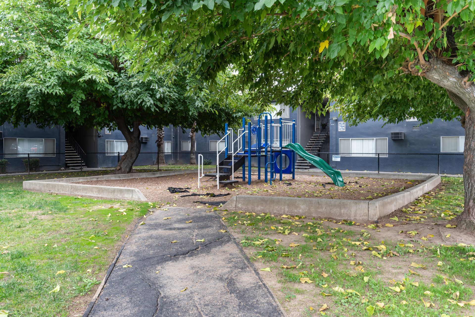 Playground nestled between trees, a paved path leading to the play structure. Green slide, blue features.