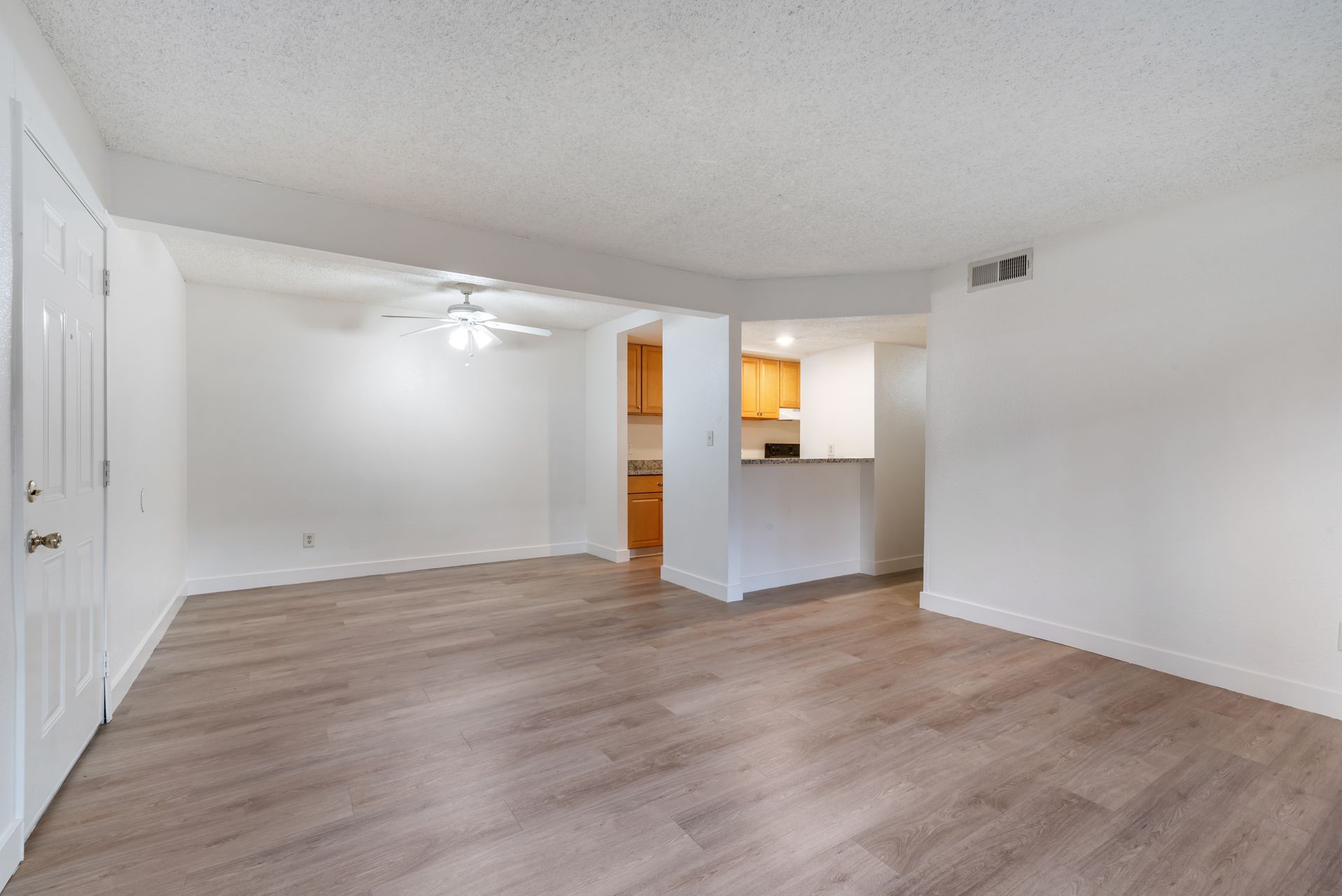 Empty living room with light wooden floors, white walls, and a view into the kitchen.