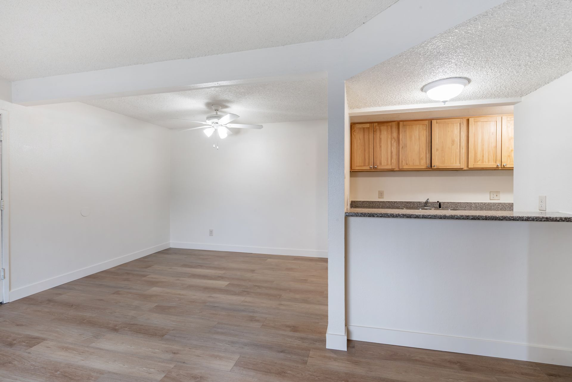 Empty apartment interior with light wood flooring, white walls, and a view into the kitchen with wooden cabinets.