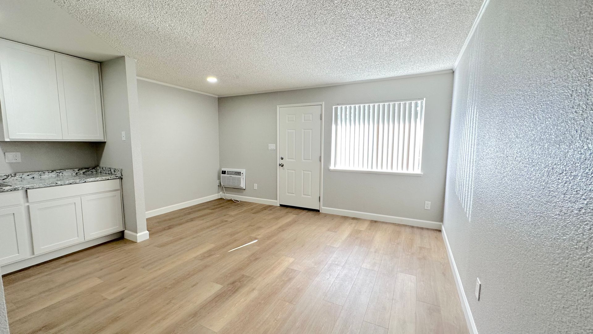 Empty apartment interior with white walls, cabinets, and door, a window with blinds, and wood-look flooring.