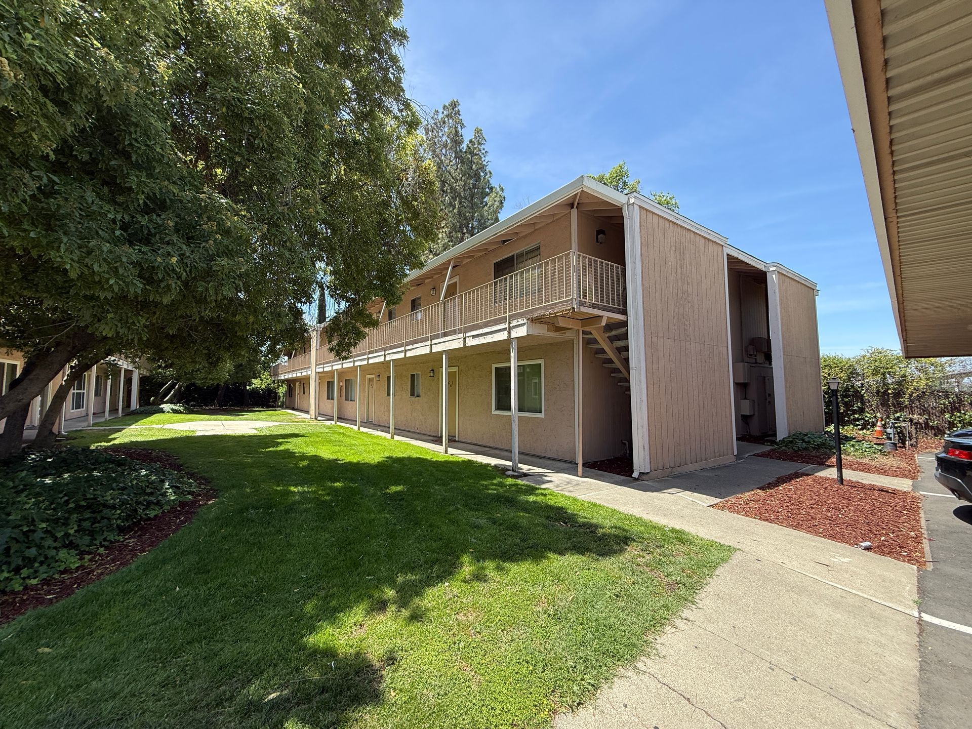 Two-story apartment building with balconies, beige siding, and green lawn, under a clear blue sky.