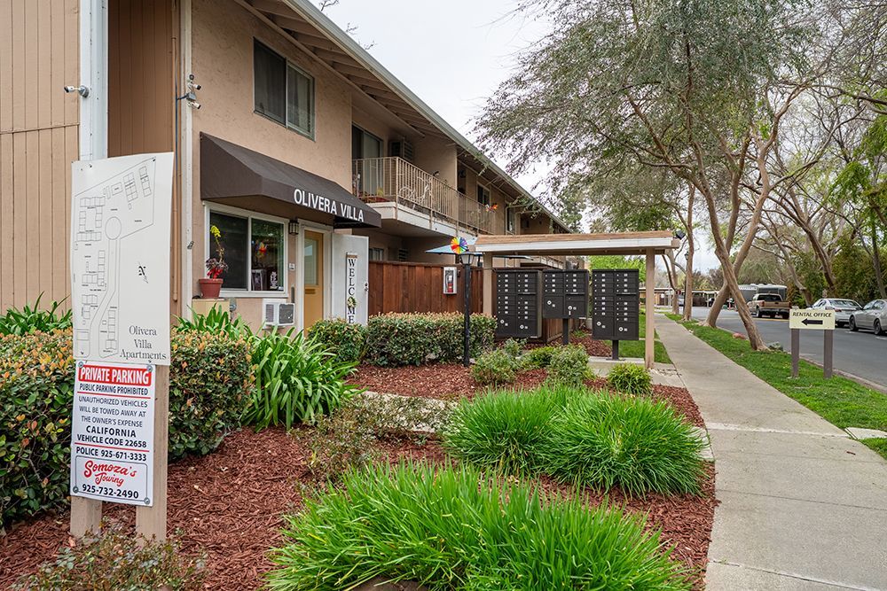 Apartment building exterior with brown siding, mailbox area, landscaping, and a sidewalk.