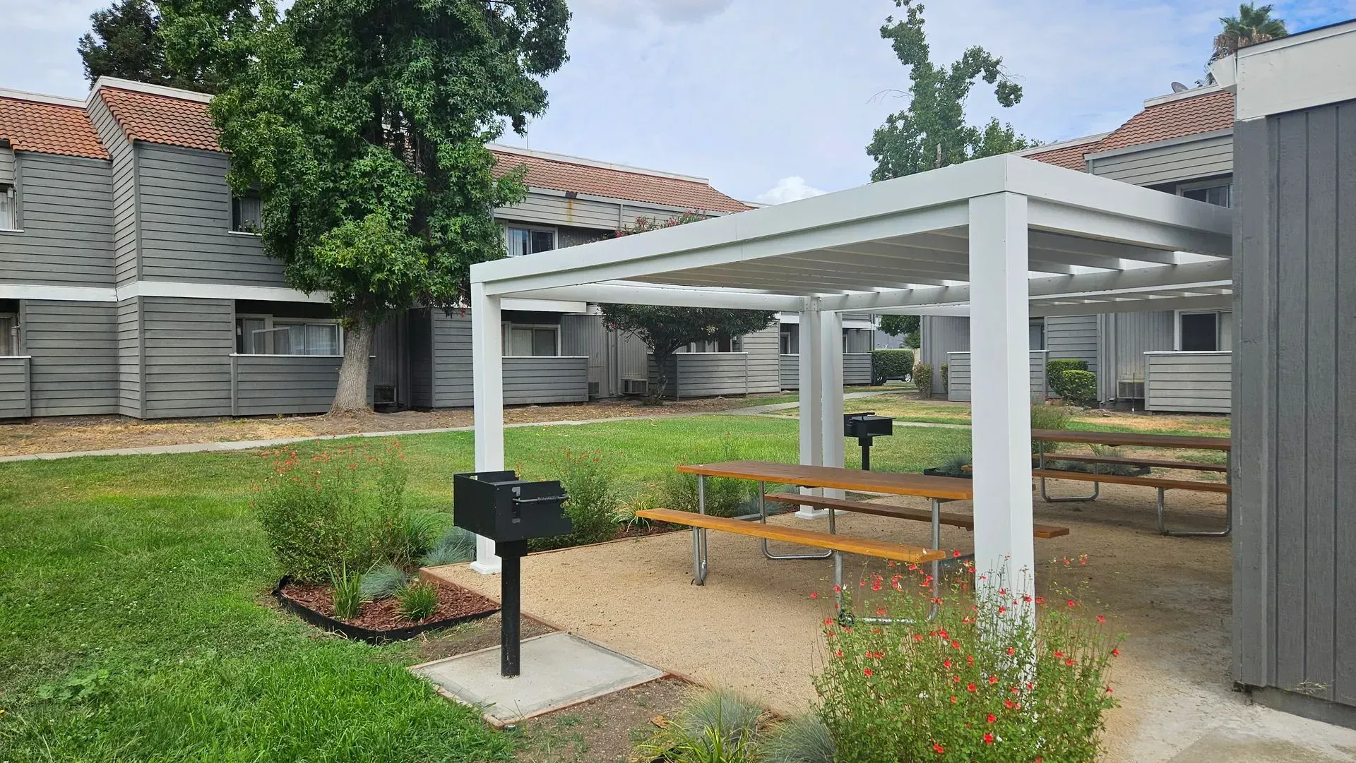 Outdoor picnic area with tables, grill, and pergola in front of apartment buildings.