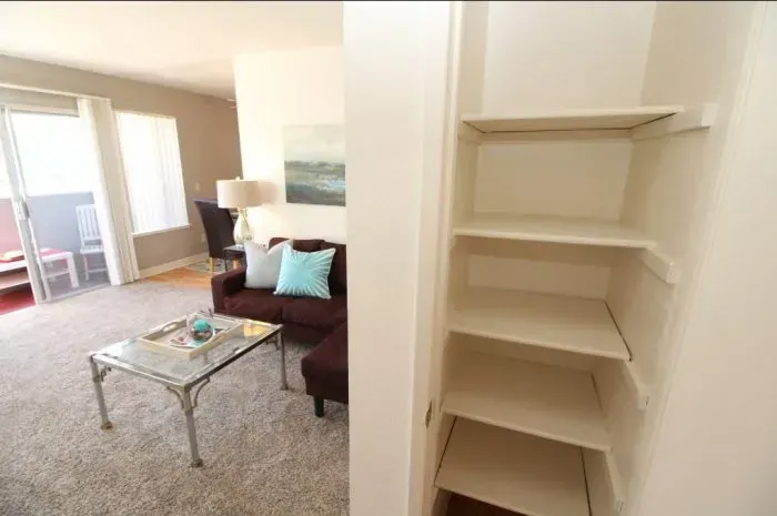 Open pantry with shelves next to a living room with a couch, table, and balcony.