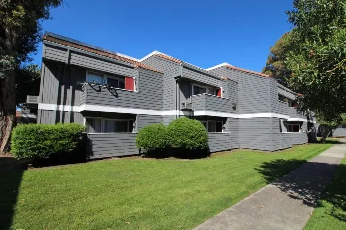 Grey apartment building with red trim, green grass, and sidewalk.