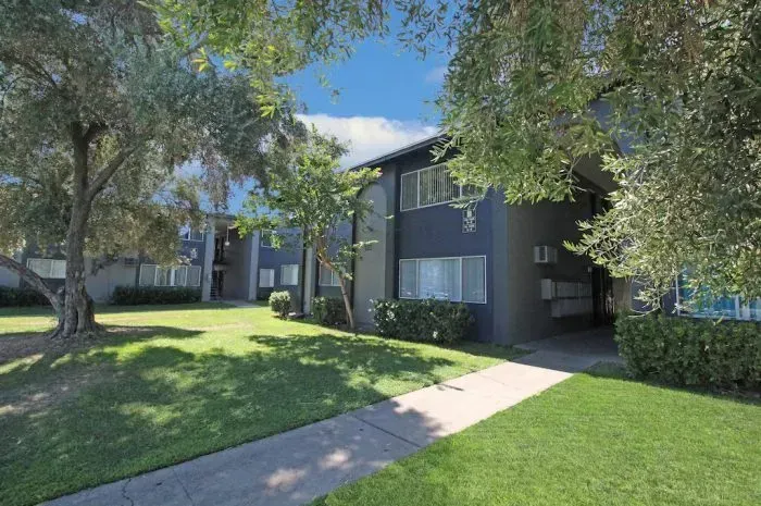 Apartment building with blue facade, green lawn, tree, and black fence. Palm trees in background.